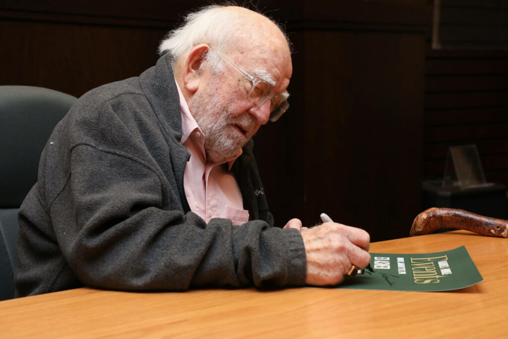 Ed Asner signing green poster wearing gray jacket pink shirt warm lighting documentary style Wallpaper