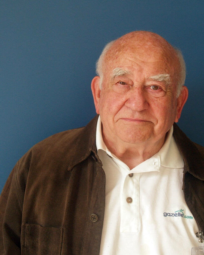 Ed Asner in brown jacket white polo shirt calm expression against plain blue wallpaper background