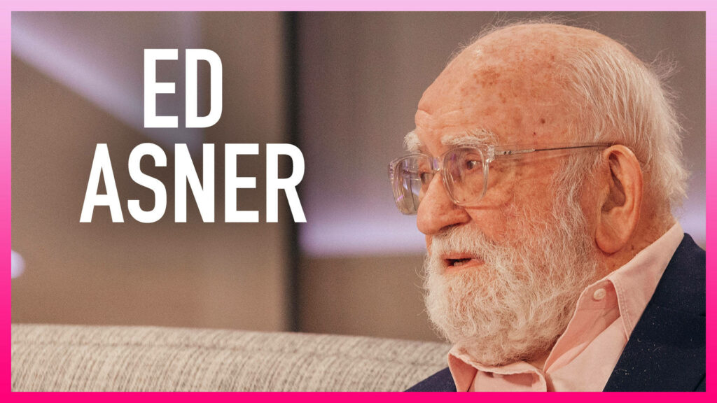 Ed Asner seated on sofa in pink shirt dark jacket thoughtful expression vibrant pink framed wallpaper