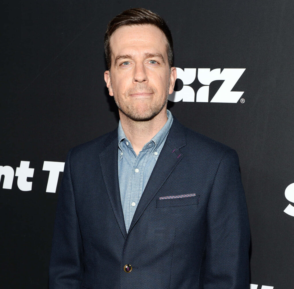 Ed Helms in navy blazer and blue shirt against dark backdrop with bold white text wallpaper