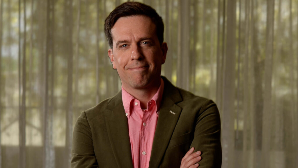 Ed Helms in green blazer and pink shirt stands confidently with crossed arms against soft curtain background Wallpaper