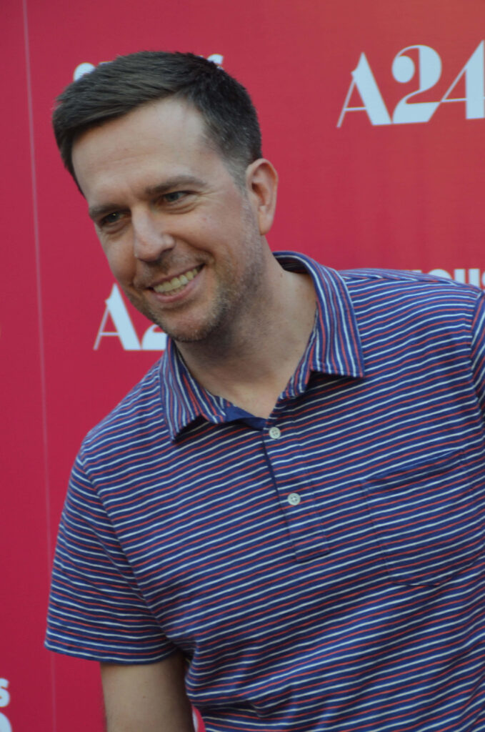 Ed Helms smiling in striped polo shirt against vibrant red A24 backdrop wallpaper background