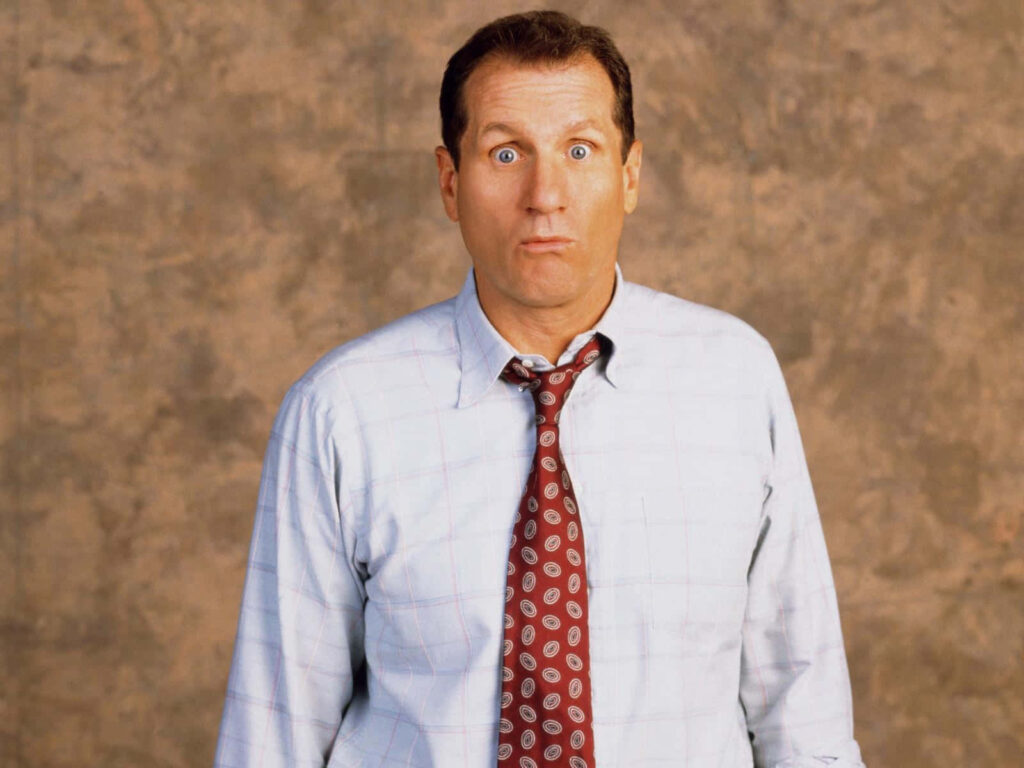 Ed ONeill portrait wearing pale blue shirt red patterned tie classic comedic expression brown background Wallpaper