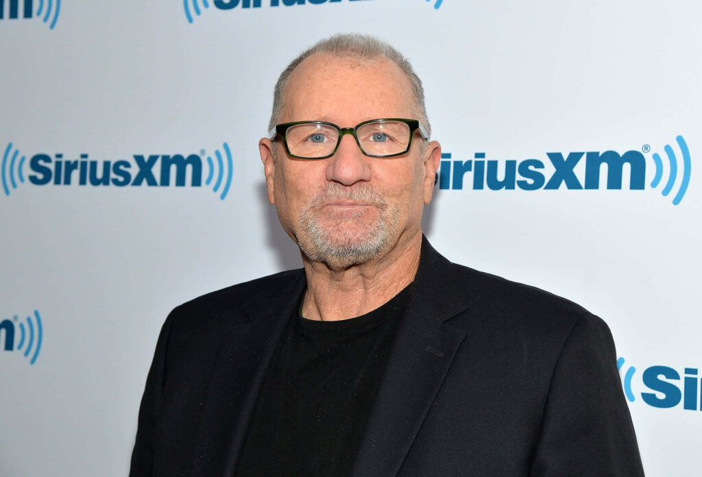 Ed ONeill in black jacket and glasses against white backdrop with SiriusXM logos wallpaper background