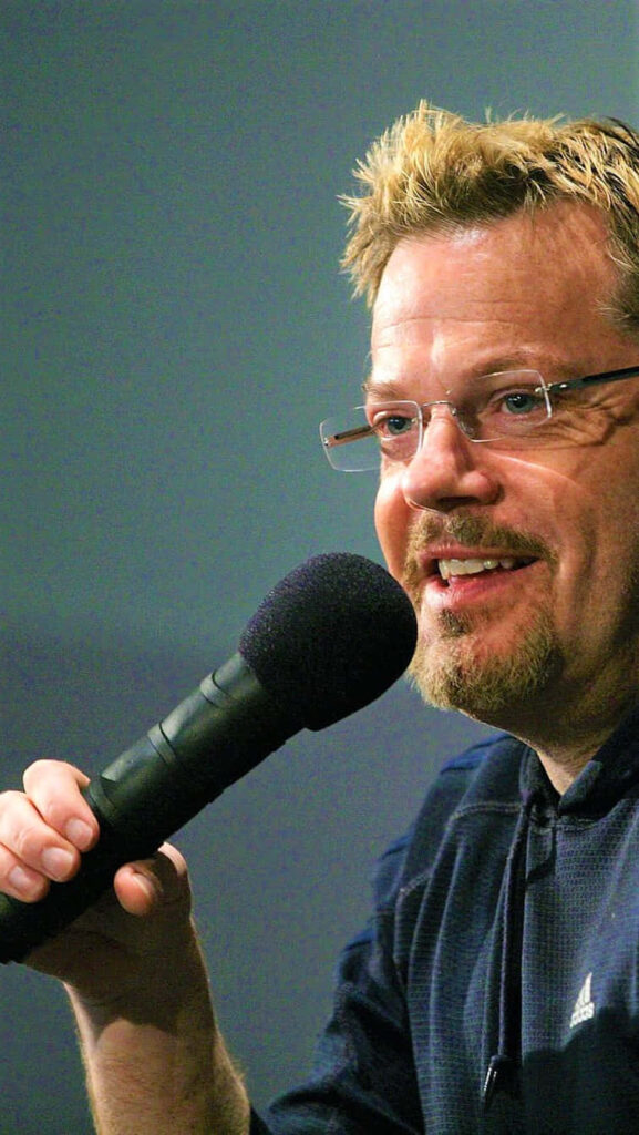 Eddie Izzard mid-performance holding microphone expressive face casual attire stage presence Wallpaper