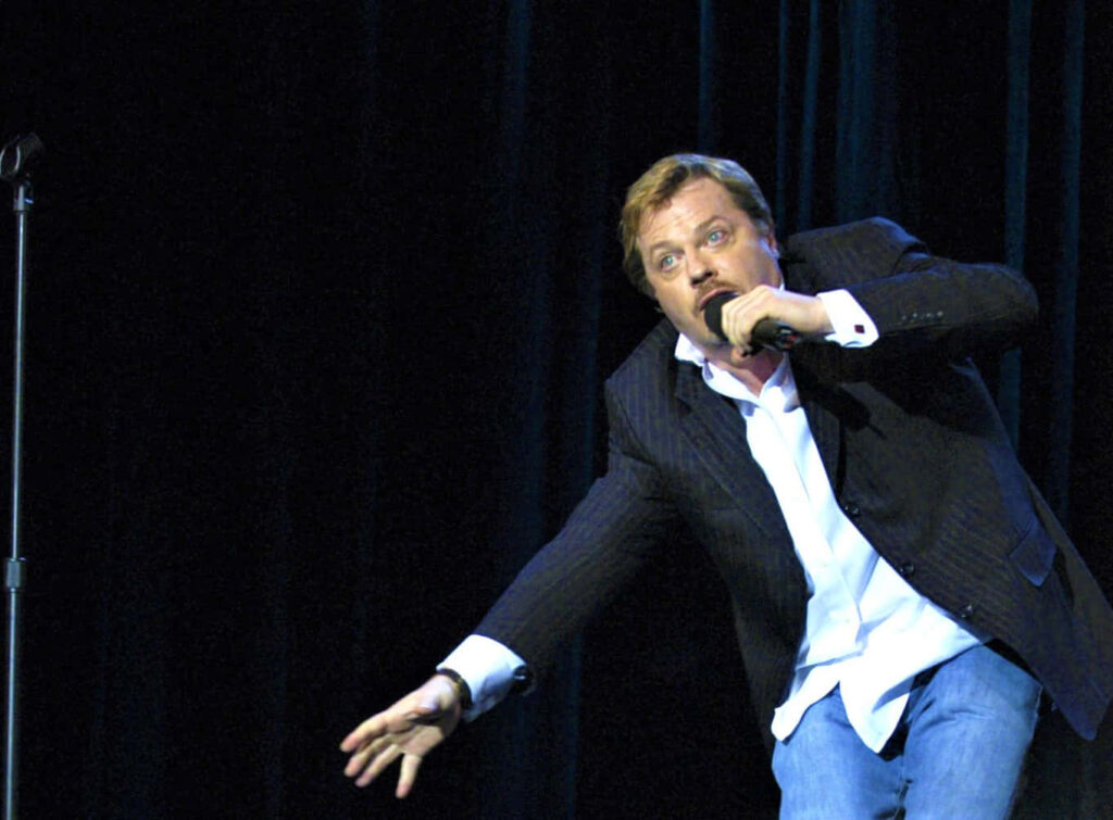 Eddie Izzard dynamic stage performance expressive gestures blue curtain theatrical comedy wallpaper