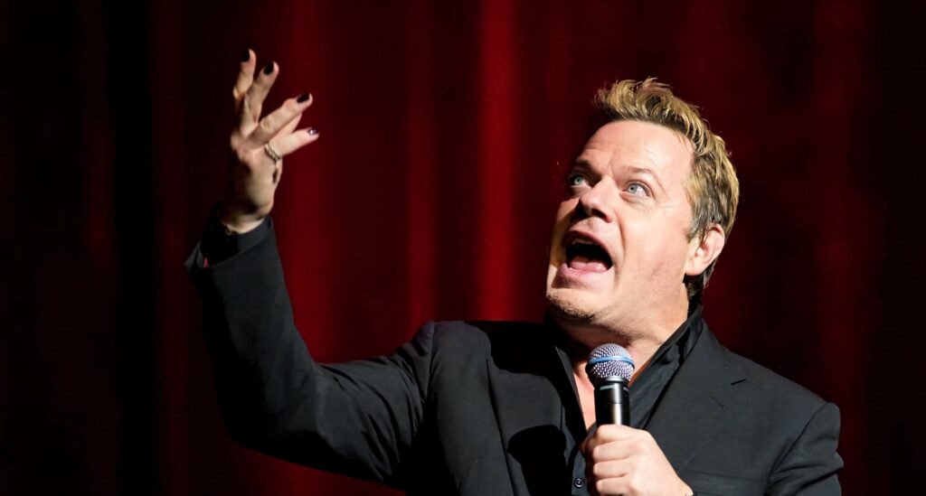 Eddie Izzard energetic stand-up comedy performance on stage vibrant red curtain backdrop wallpaper