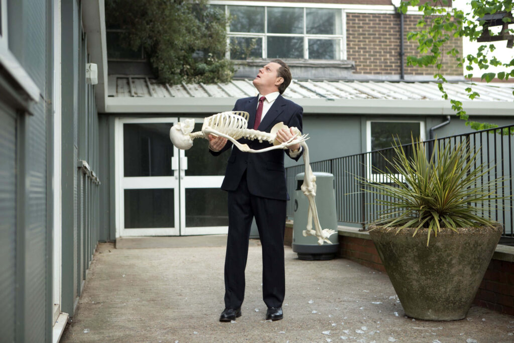 Eddie Izzard in formal suit holding human skeleton outside modern building with puzzled expression Wallpaper