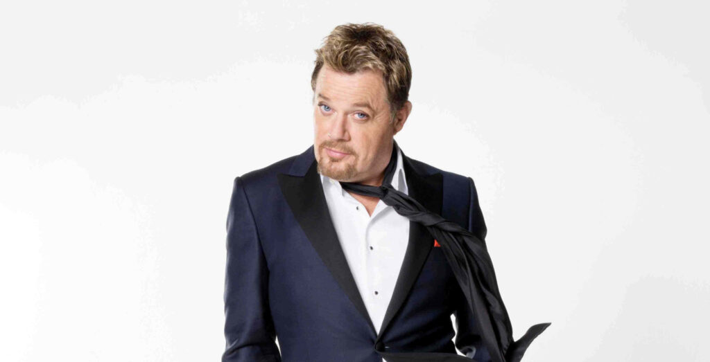 Eddie Izzard in sharp dark suit with moving black tie on light background high resolution wallpaper