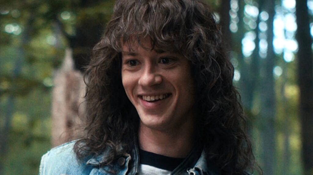 Eddie Munson smiling outdoors with curly hair denim jacket lush greenery vibrant wallpaper background