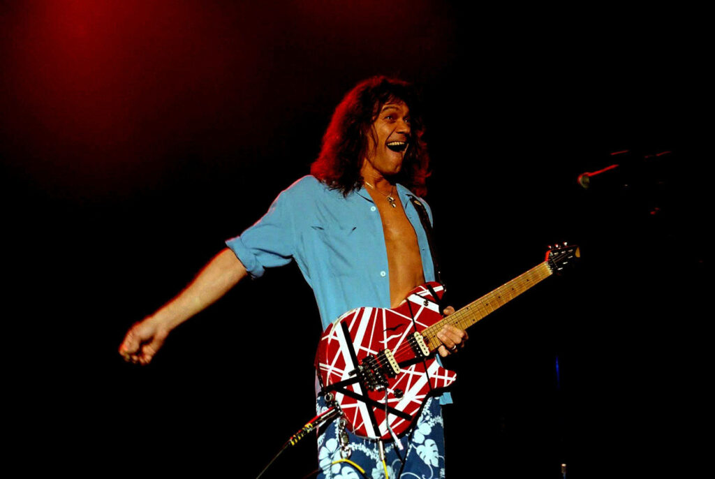 Eddie Van Halen playing iconic striped guitar smiling on stage with vibrant red yellow lights Wallpaper