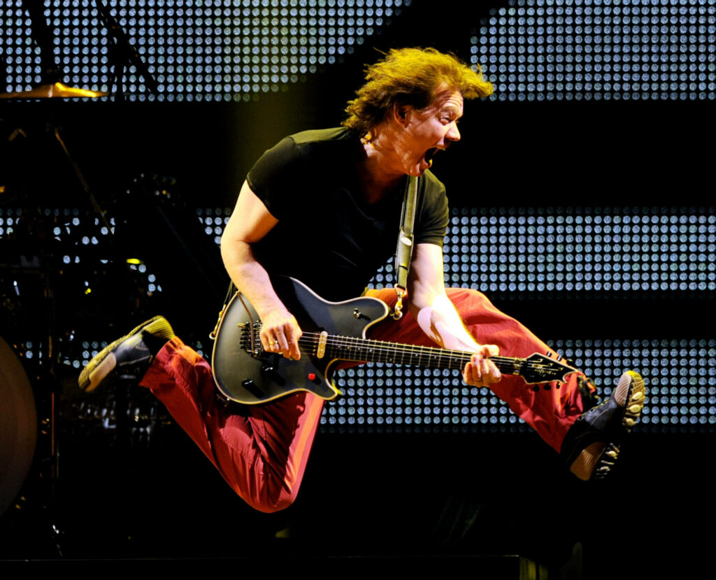 Eddie Van Halen mid-air guitar jump glowing stage lights red pants energetic rock performance Wallpaper
