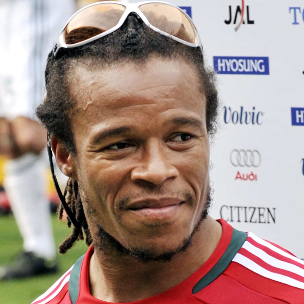Closeup headshot of Edgar Davids with signature dreadlocks and goggles set against blurred logos background Wallpaper