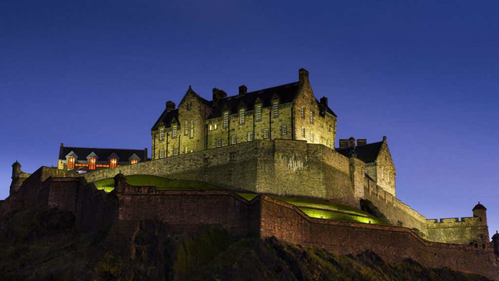 Edinburgh Castle skyline at night glowing golden on rugged stone with deep blue sky background Wallpaper