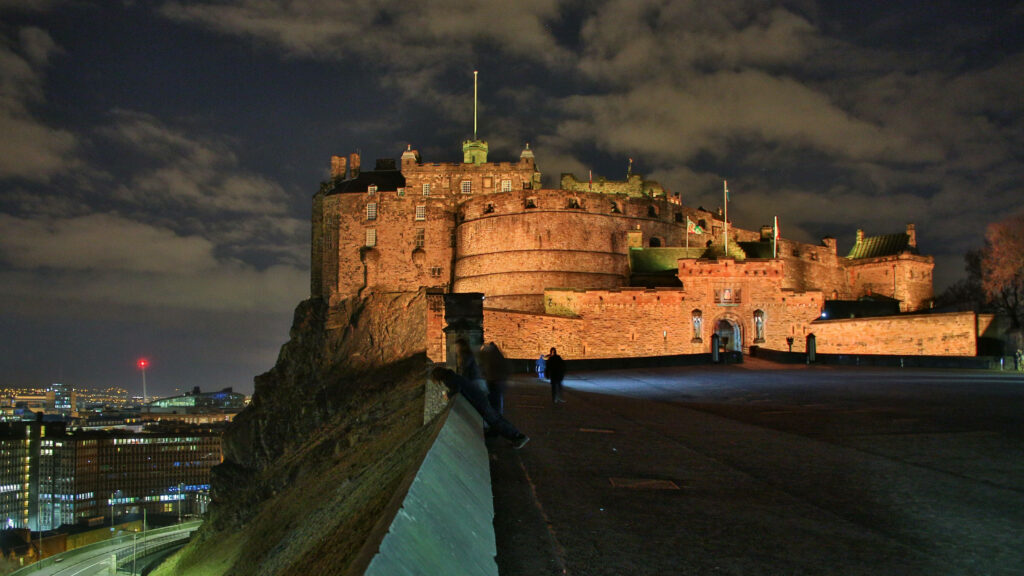 Edinburgh Castle entrance at night illuminated by golden lights under a dramatic cloudy sky Wallpaper