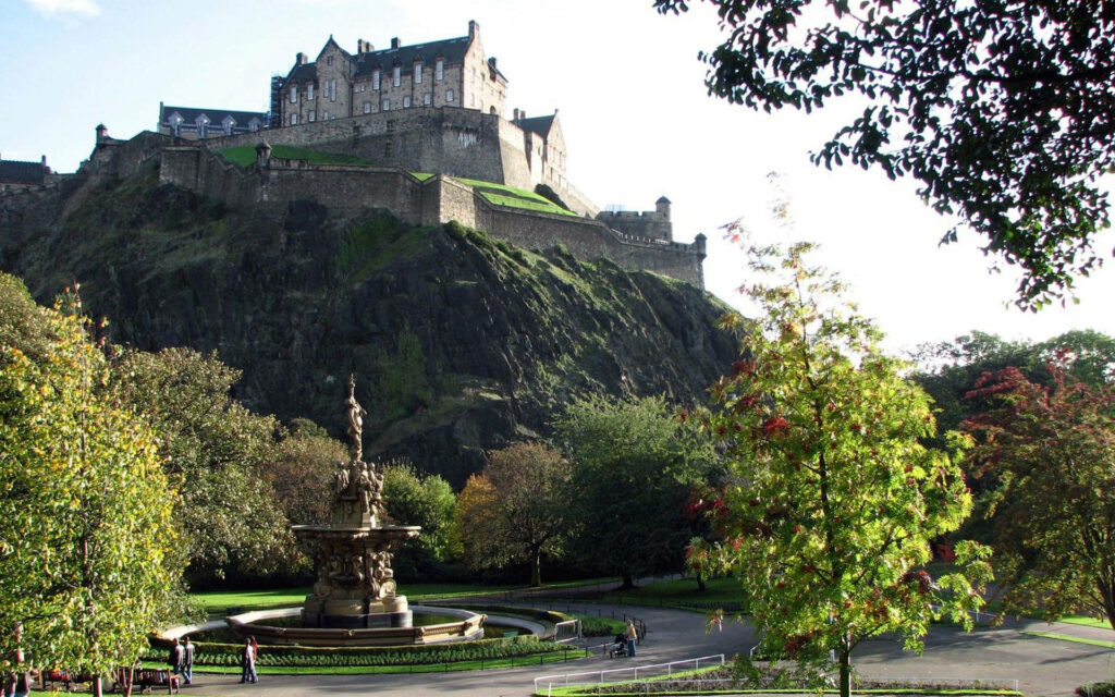 Lush park with classical fountain under historic Edinburgh Castle on rocky hill in sunlight Wallpaper