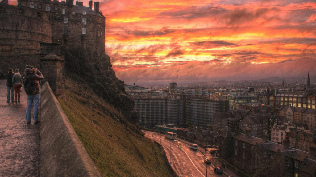 Tourists walking ancient stone path near Edinburgh Castle at sunset with city and vibrant sky views Wallpaper