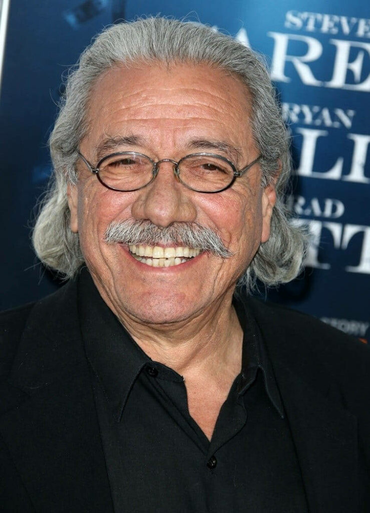 Edward James Olmos smiling with gray hair mustache and glasses against blue backdrop wallpaper
