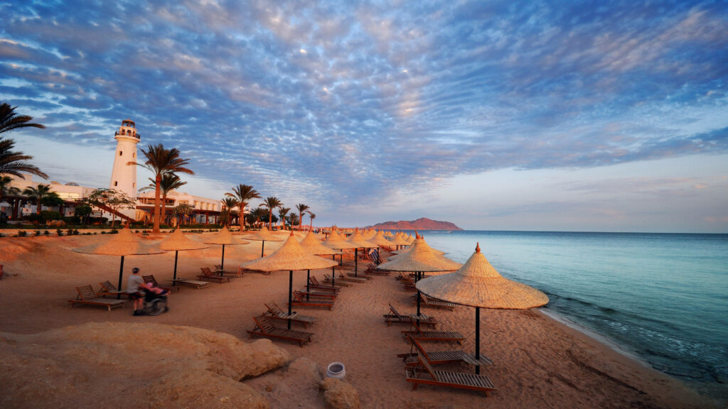 Serene Egypt beach sunset with palm trees thatched umbrellas wooden loungers and lighthouse Wallpaper