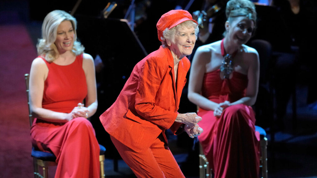 Elaine Stritch in red suit performing on stage with women in red dresses under theater lights Wallpaper
