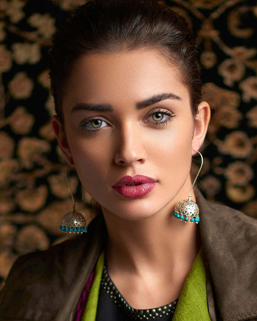 Elegant portrait of Amy Jackson with intricate jewelry and rich patterned wallpaper background