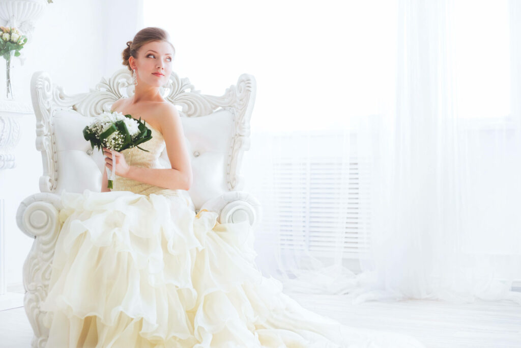 Elegant bride in ruffled ivory gown seated on ornate white chair holding bouquet in bright room Wallpaper