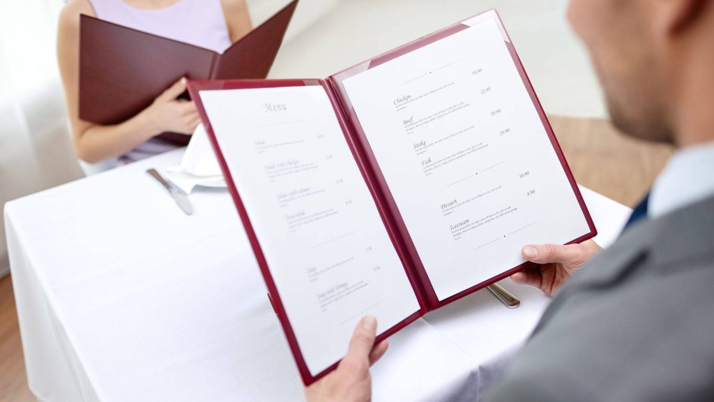 Couple dining at restaurant holding red menus seated at white tablecloth table bright elegant setting Wallpaper