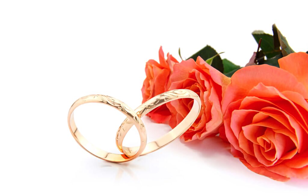 Two thin gold band engagement rings with engraved details beside vibrant orange roses on white Wallpaper