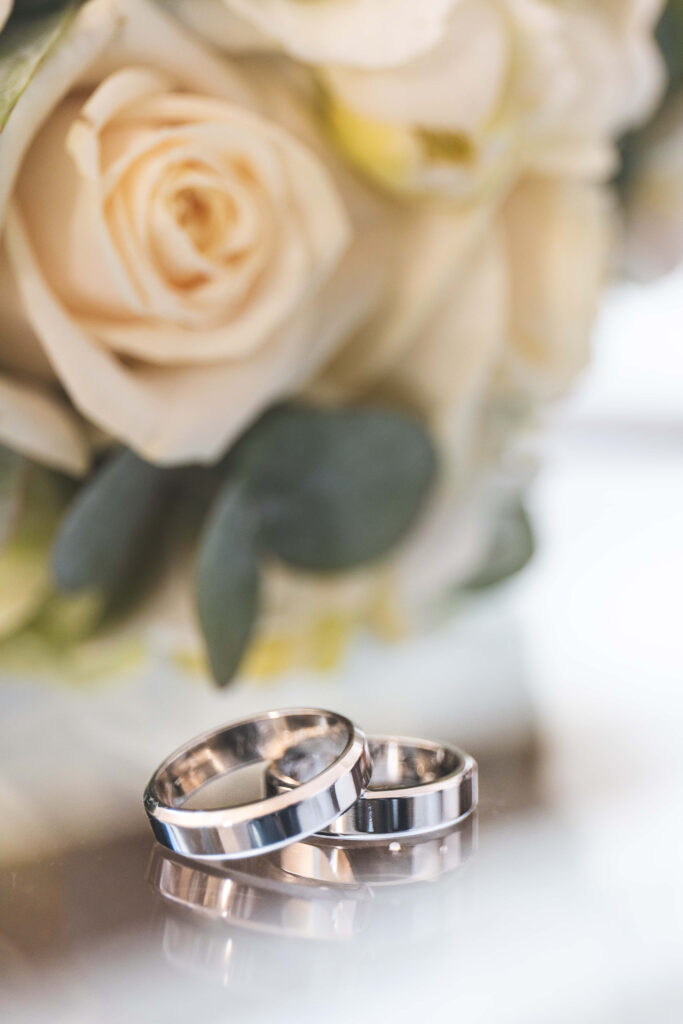 Elegant silver band engagement rings on reflective surface with white roses and greenery background Wallpaper