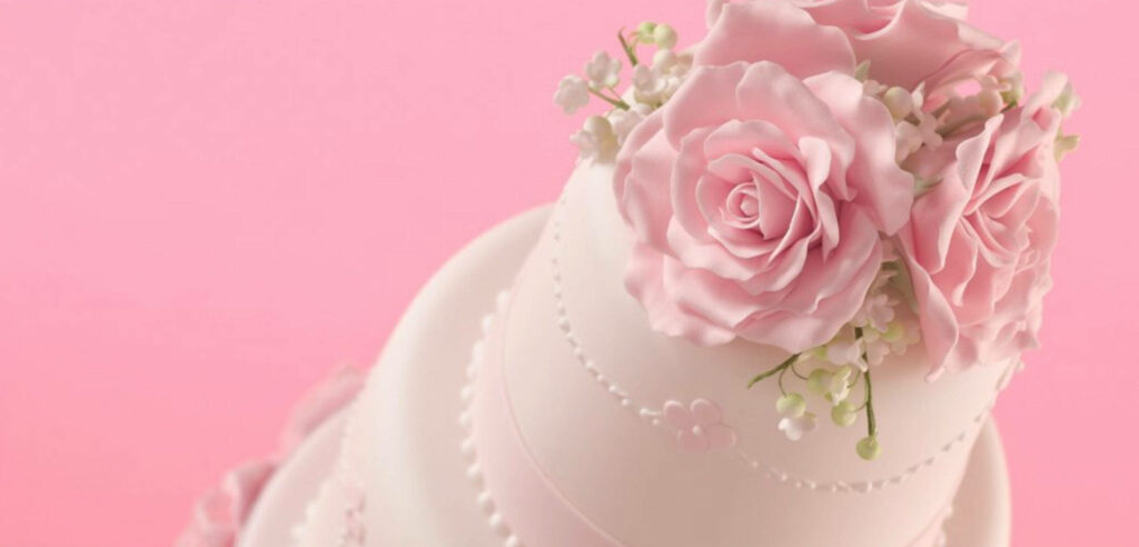 Elegant white wedding cake with real pink roses and floral accents on soft pink background Wallpaper