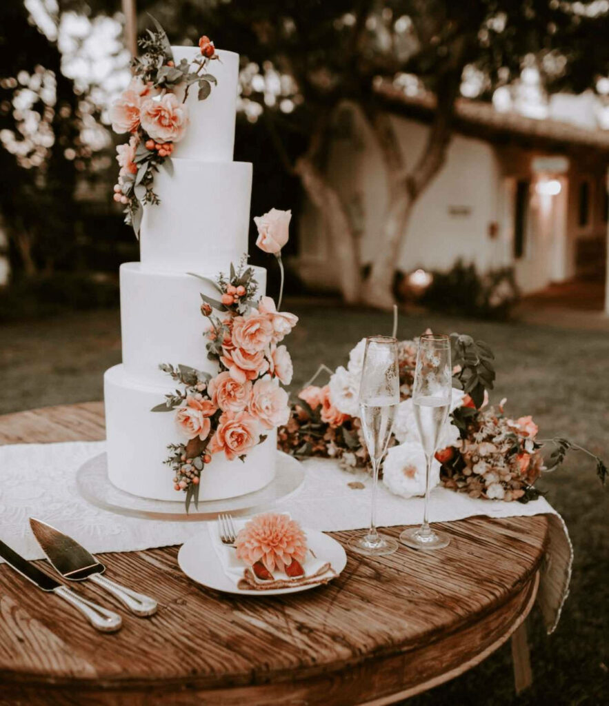 Elegant white wedding cake with pink roses and greenery on rustic wooden table outdoors Wallpaper
