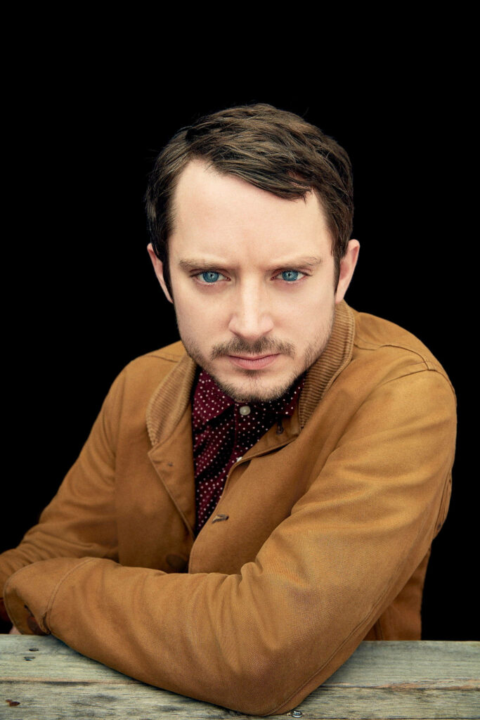 Elijah Wood portrait with intense blue eyes brown jacket dotted shirt minimalist black background Wallpaper