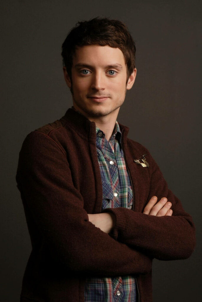 Elijah Wood studio portrait with arms crossed wearing plaid shirt and maroon jacket on dark background Wallpaper
