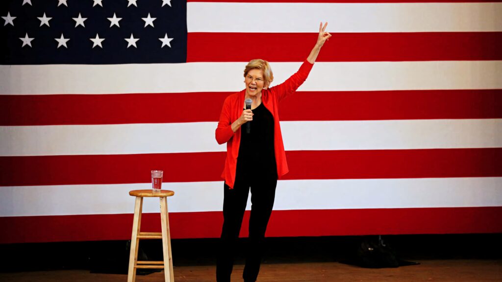 Elizabeth Warren in red jacket making peace sign with microphone and American flag backdrop Wallpaper