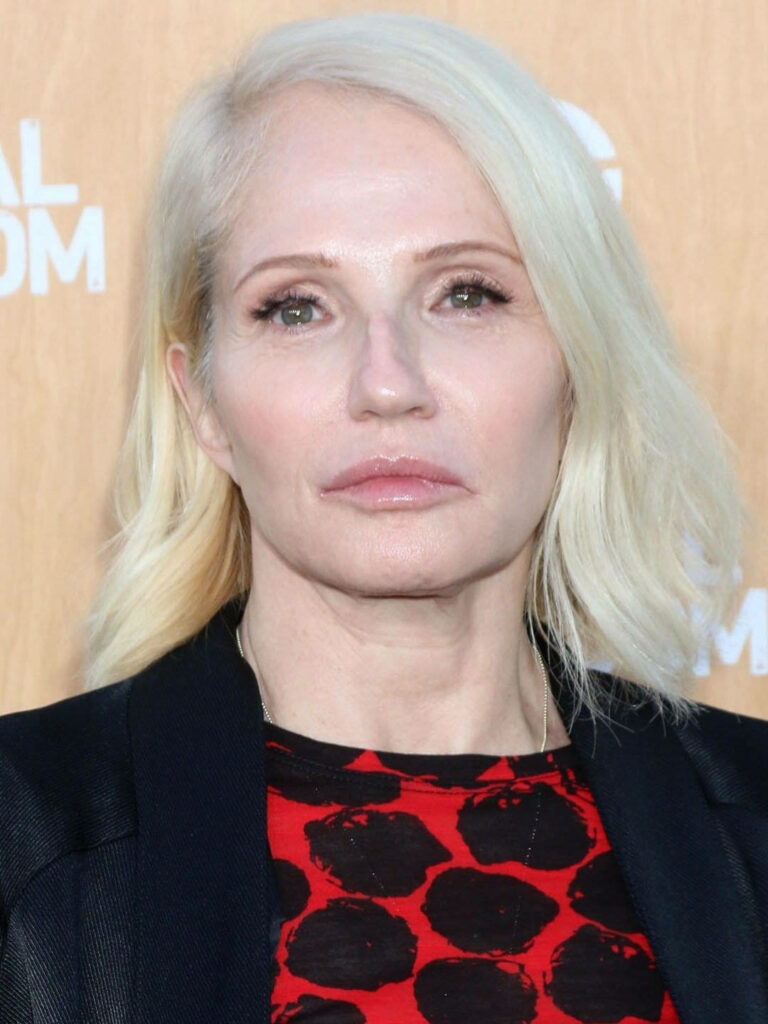Ellen Barkin in red black patterned top and black blazer against warm textured background wallpaper