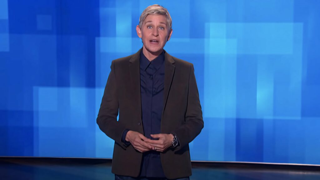 Ellen DeGeneres on The Ellen Show set with vibrant blue geometric wallpaper background