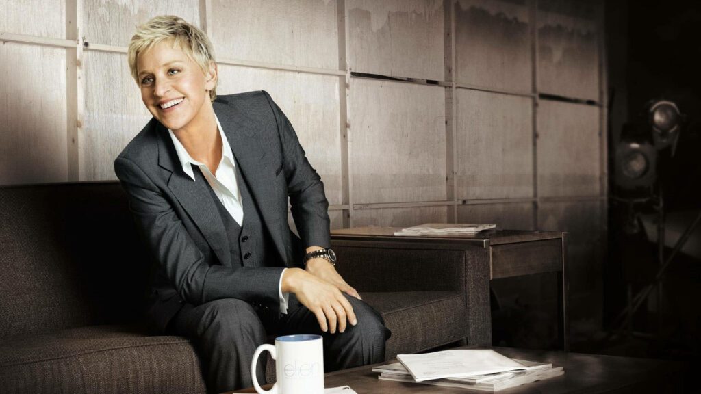 Ellen DeGeneres smiling in dark grey satin suit sitting on sofa in stylish relaxed studio setting Wallpaper