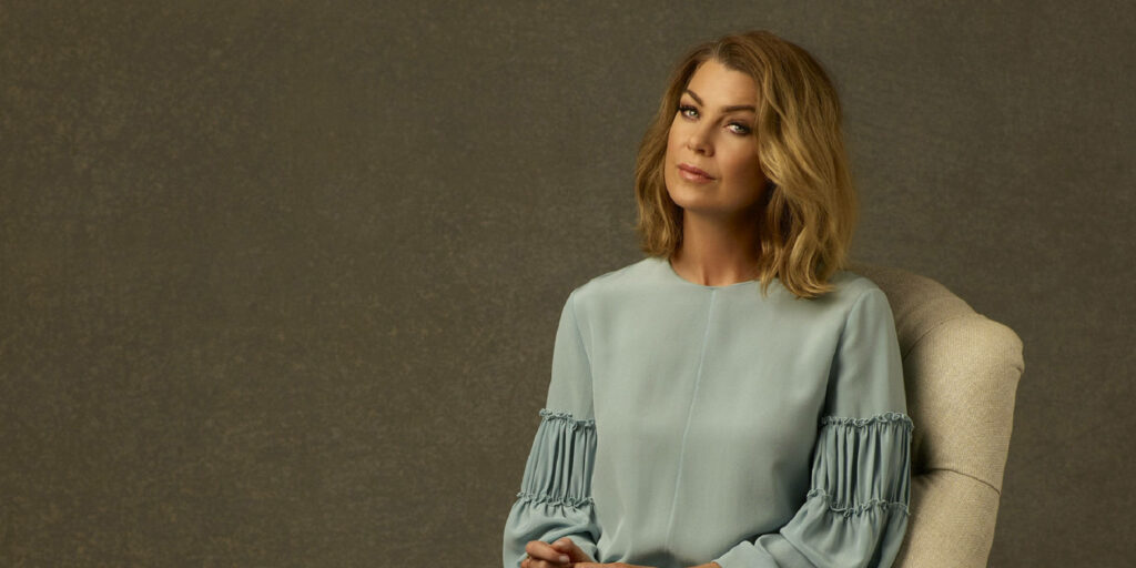 Ellen Pompeo seated in light blue ruffle sleeve blouse against textured neutral wallpaper background