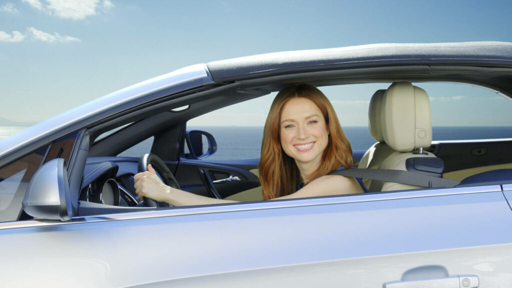 Ellie Kemper smiling driving silver Buick with beige interior ocean and blue sky background wallpaper