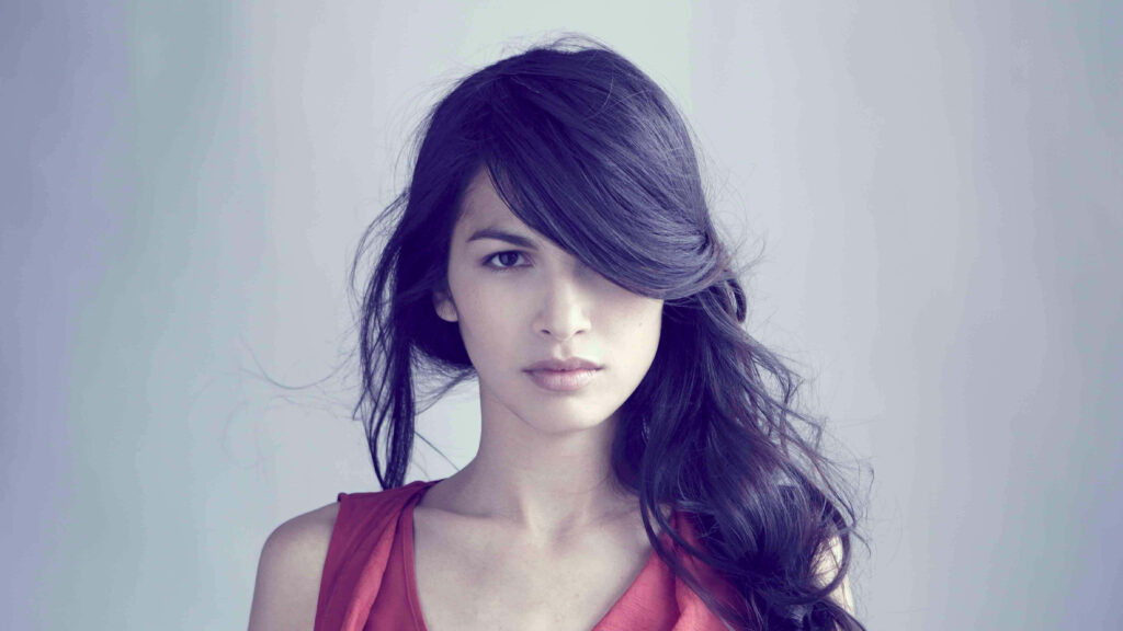 Elodie Yung in vibrant red top with dark wavy hair softly lit against pale minimalist background Wallpaper