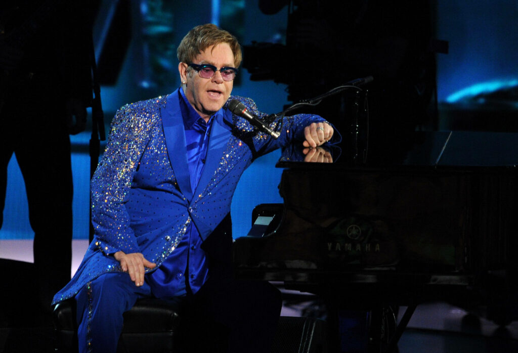 Elton John performing piano in blue rhinestone suit on dark stage showcasing vibrant energy Wallpaper
