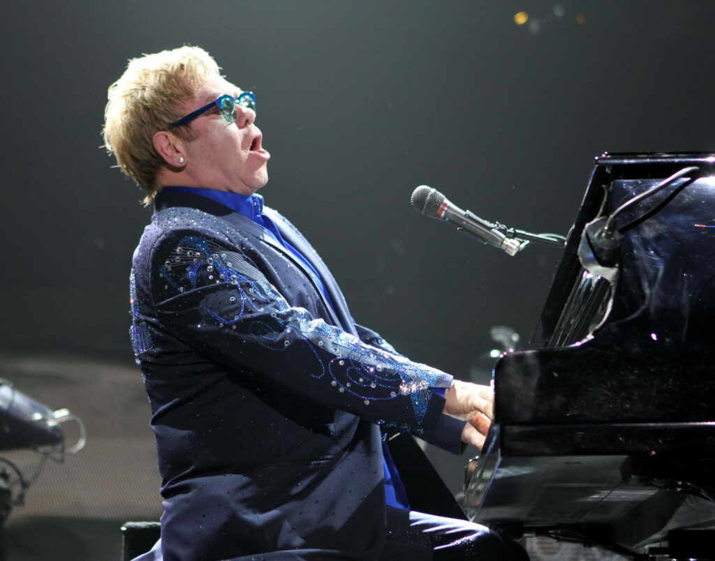 Elton John performing live at piano in glittering blue suit with iconic sunglasses and stage lights Wallpaper