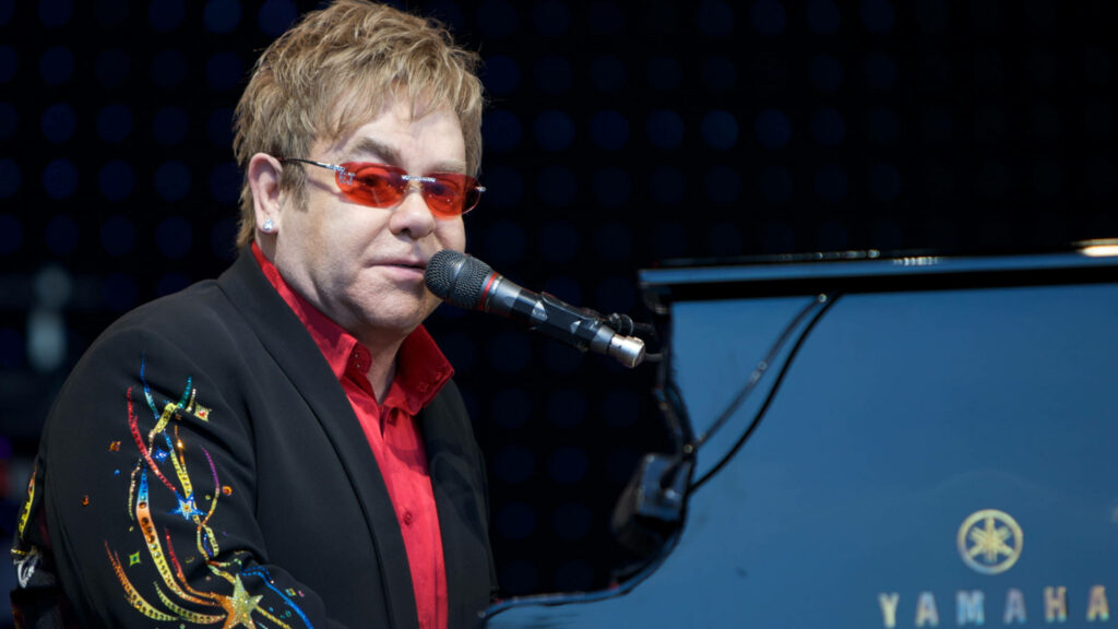 Elton John performing live at piano wearing black embroidered jacket red shirt and signature glasses Wallpaper