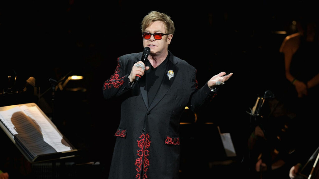 Elton John on stage in dark coat with red patterns and round red glasses live concert wallpaper
