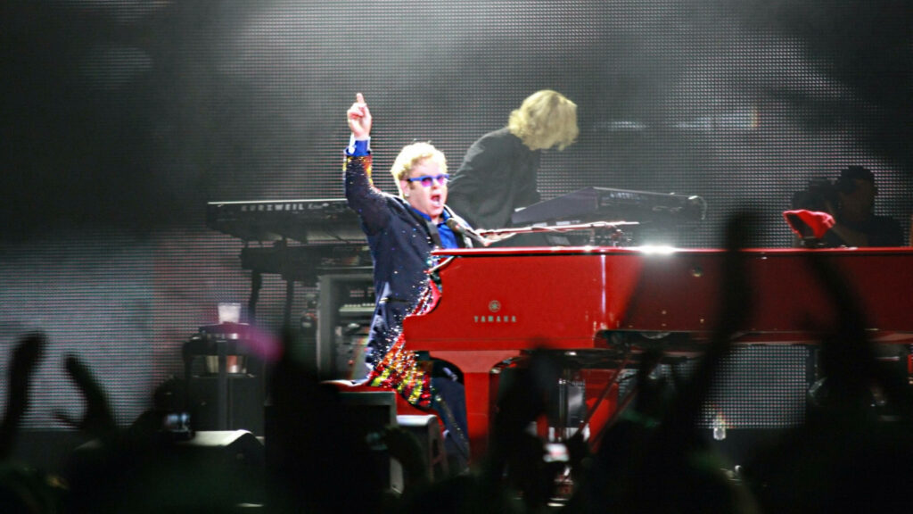 Elton John performing live at a vibrant red piano in colorful suit and blue glasses onstage Wallpaper
