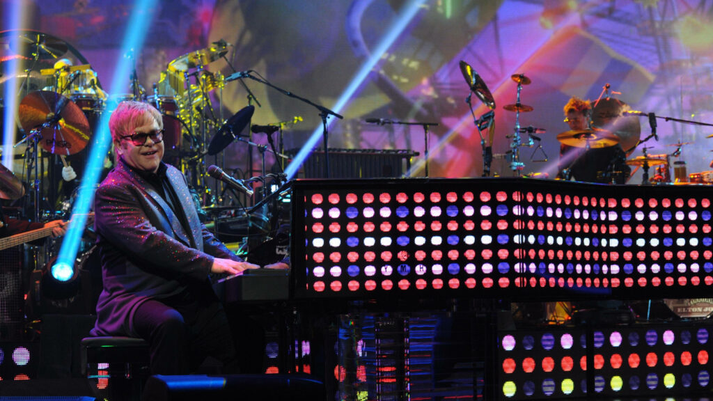 Elton John performing live on stage with vibrant illuminated piano and colorful dynamic lights Wallpaper