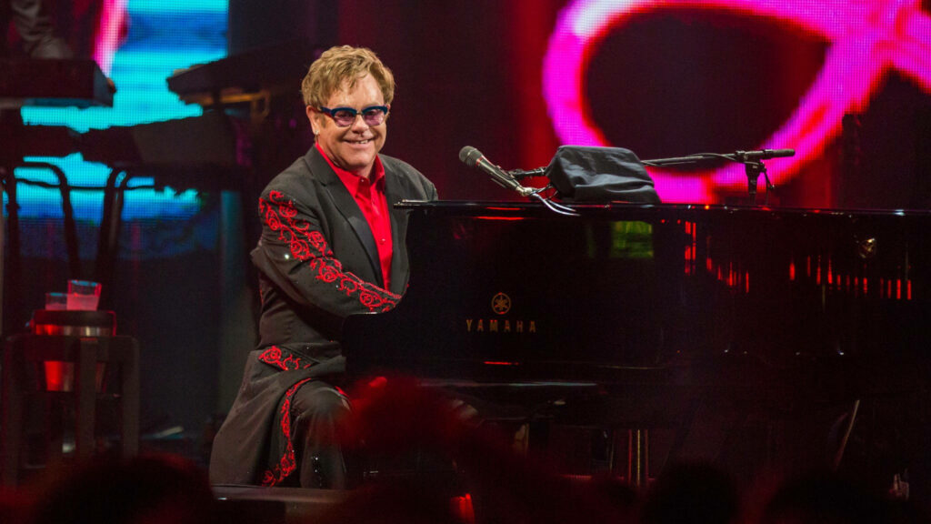 Elton John performing live at grand piano wearing red black suit colorful energetic concert background Wallpaper