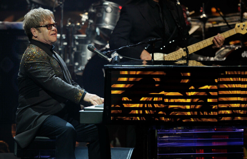 Elton John performing live on stage playing grand piano in sparkling suit with golden lights Wallpaper