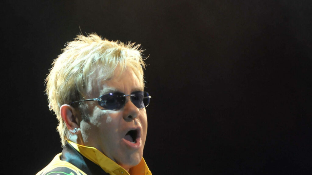 Elton John mid-performance with blond hair bold sunglasses vibrant lighting dark stage background Wallpaper