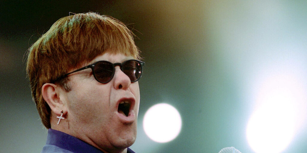 Elton John mid-performance wearing round sunglasses and cross earring vibrant concert wallpaper